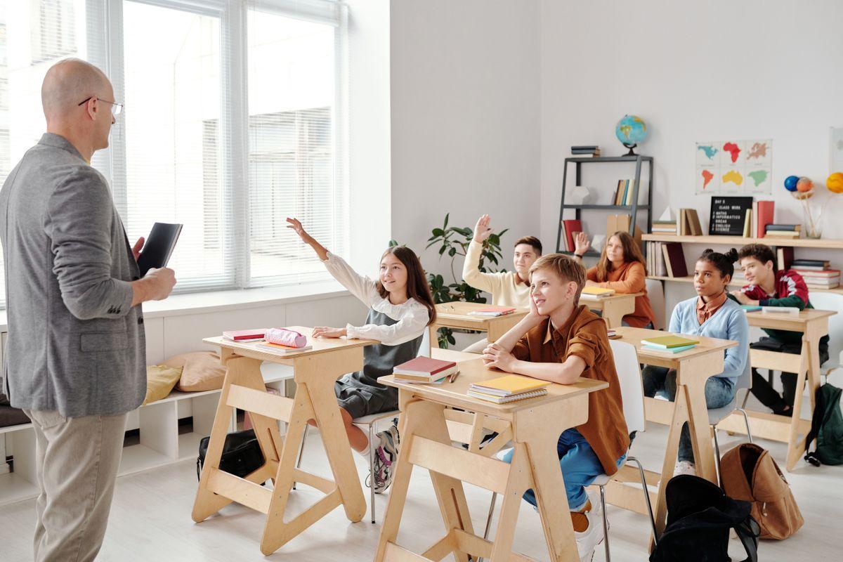 A teacher in a bright classroom with students raising their hands